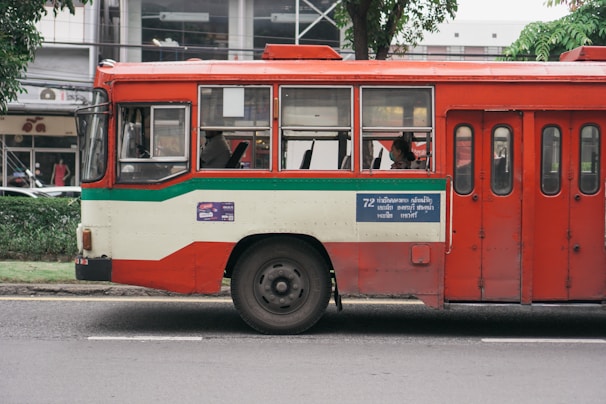 A red and cream bus parked on a street, with a green stripe running along its side. There are passengers visible through the windows, and a blue sign with text is displayed on the side of the bus. Trees and a building can be seen in the background.