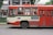 A red and cream bus parked on a street, with a green stripe running along its side. There are passengers visible through the windows, and a blue sign with text is displayed on the side of the bus. Trees and a building can be seen in the background.