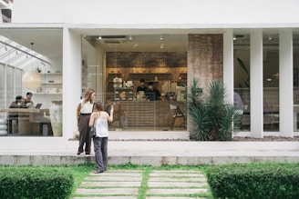 A modern cafe with large glass windows showcasing a minimalist interior. Two women stand outside near the entrance, one taking a photo, while inside, people are sitting at tables, working or engaging in conversation. The decor includes large planters and a clean, neutral color scheme providing a calming atmosphere.