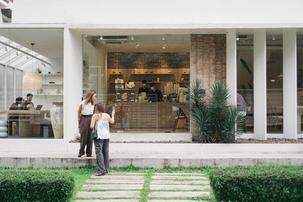 A modern cafe with large glass windows showcasing a minimalist interior. Two women stand outside near the entrance, one taking a photo, while inside, people are sitting at tables, working or engaging in conversation. The decor includes large planters and a clean, neutral color scheme providing a calming atmosphere.