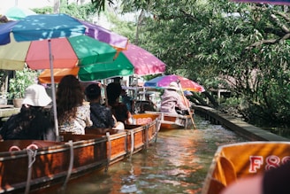 A group of people seated in wooden boats with colorful umbrellas, navigating a narrow canal surrounded by lush greenery and trees.