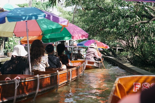 A group of people seated in wooden boats with colorful umbrellas, navigating a narrow canal surrounded by lush greenery and trees.