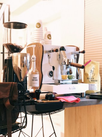 A well-organized coffee station features a sleek espresso machine with wooden accents, accompanied by a coffee grinder filled with beans. A bottle of vanilla syrup and a yellow espresso cup add visual interest. Additionally, a stack of disposable coffee cups with a logo is neatly arranged, and a bag of coffee beans is positioned on the right. A red cloth and brown apron are draped over a black wire chair, hinting at a cozy, caf&eacute;-like atmosphere.