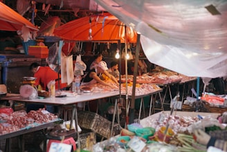 A bustling market scene with vendors and raw meat on display under orange umbrellas. People are busy arranging meat on tables, with plastic containers and various items around. Some stall lights hang overhead, illuminating the area.