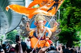 A large, colorful statue of the Hindu deity Ganesha with intricate details is being paraded through a bustling crowd. The statue is adorned with various ornaments and jewelry, and it is placed on a decorative pedestal. An orange flag with black symbols flies prominently in the foreground. The setting is urban, with a modern structure visible on the left and lush green trees on the right. Many people are gathered around, some capturing the moment with their phones.