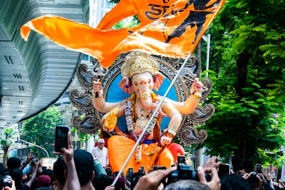 A large, colorful statue of the Hindu deity Ganesha with intricate details is being paraded through a bustling crowd. The statue is adorned with various ornaments and jewelry, and it is placed on a decorative pedestal. An orange flag with black symbols flies prominently in the foreground. The setting is urban, with a modern structure visible on the left and lush green trees on the right. Many people are gathered around, some capturing the moment with their phones.