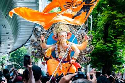 A large, colorful statue of the Hindu deity Ganesha with intricate details is being paraded through a bustling crowd. The statue is adorned with various ornaments and jewelry, and it is placed on a decorative pedestal. An orange flag with black symbols flies prominently in the foreground. The setting is urban, with a modern structure visible on the left and lush green trees on the right. Many people are gathered around, some capturing the moment with their phones.