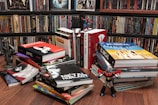 A room with shelves filled with colorful DVDs and a variety of large illustrated books stacked on the floor. Prominent books include 'Persepolis' and 'El Incal'. Small toy figures are placed between the books, including a Spiderman and a robot-like figure. The setting appears to be vibrant and eclectic.