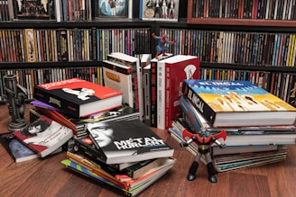 A room with shelves filled with colorful DVDs and a variety of large illustrated books stacked on the floor. Prominent books include 'Persepolis' and 'El Incal'. Small toy figures are placed between the books, including a Spiderman and a robot-like figure. The setting appears to be vibrant and eclectic.