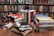 A room with shelves filled with colorful DVDs and a variety of large illustrated books stacked on the floor. Prominent books include 'Persepolis' and 'El Incal'. Small toy figures are placed between the books, including a Spiderman and a robot-like figure. The setting appears to be vibrant and eclectic.