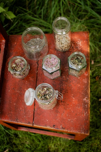 Several glass jars containing different types of dried herbs are arranged on a weathered red wooden table. The collection of jars varies in size and shape, with some open and others closed, showcasing a variety of colorful herbs and seeds.