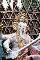 A statue of Lord Ganesha, characterized by an elaborate headdress and intricate jewelry. The figure holds a stringed musical instrument in a decorative pose. The background features ornate triangular patterns with detailed gold and red designs.