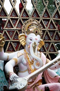 A statue of Lord Ganesha, characterized by an elaborate headdress and intricate jewelry. The figure holds a stringed musical instrument in a decorative pose. The background features ornate triangular patterns with detailed gold and red designs.