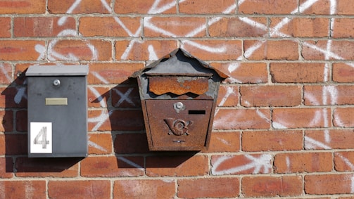 Two mailboxes are mounted on a brick wall covered in graffiti. One mailbox is modern and gray, with the number 4 displayed. The other is old and rusted, with a decorative emblem on the front.