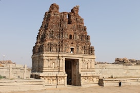 A historic stone temple structure with intricate carvings and a weathered appearance, set in an open area with stone walls and rocky terrain surrounding it. The temple features multiple tiers and detailed architectural elements typical of ancient Indian temple architecture.