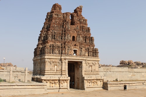A historic stone temple structure with intricate carvings and a weathered appearance, set in an open area with stone walls and rocky terrain surrounding it. The temple features multiple tiers and detailed architectural elements typical of ancient Indian temple architecture.