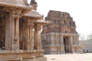 An ancient temple structure featuring intricately carved stone pillars and a central shrine with a tall gopuram (tower). The architecture includes detailed engravings and sculptures typical of historic South Indian temple design. The area is illuminated by natural light, highlighting the texture of the stonework.