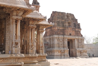 An ancient temple structure featuring intricately carved stone pillars and a central shrine with a tall gopuram (tower). The architecture includes detailed engravings and sculptures typical of historic South Indian temple design. The area is illuminated by natural light, highlighting the texture of the stonework.