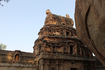 An ancient stone temple structure with intricate carvings and detailed architecture. The building is surrounded by a massive rock formation on one side and clear blue sky above. The temple's upper sections feature ornate sculptures.