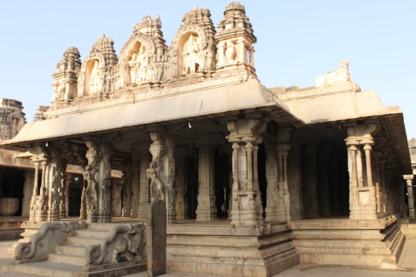 A historic temple structure with intricate carvings on the stone columns and ornate figures topping the roof. The architecture features a series of detailed pillars supporting a large, flat roof with numerous statues and embellishments.