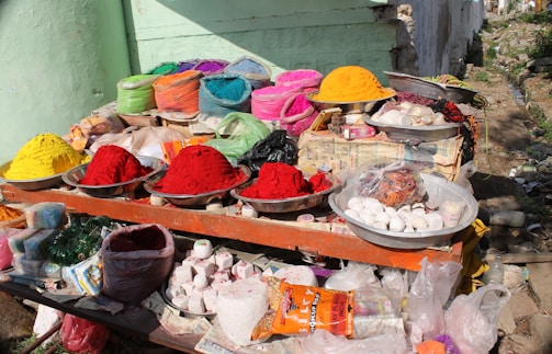 A roadside stall with a variety of colorful powdered dyes and snacks displayed in metal bowls and bags. Vivid colors like red, yellow, orange, pink, and green are prominently featured. Various products, including incense sticks and packaged items, are also visible among the array.