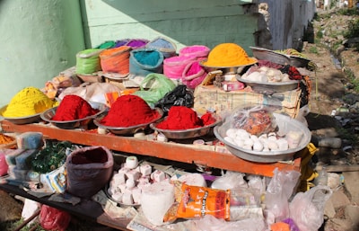 A roadside stall with a variety of colorful powdered dyes and snacks displayed in metal bowls and bags. Vivid colors like red, yellow, orange, pink, and green are prominently featured. Various products, including incense sticks and packaged items, are also visible among the array.