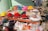 A roadside stall with a variety of colorful powdered dyes and snacks displayed in metal bowls and bags. Vivid colors like red, yellow, orange, pink, and green are prominently featured. Various products, including incense sticks and packaged items, are also visible among the array.