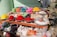 A roadside stall with a variety of colorful powdered dyes and snacks displayed in metal bowls and bags. Vivid colors like red, yellow, orange, pink, and green are prominently featured. Various products, including incense sticks and packaged items, are also visible among the array.