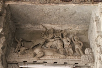 A detailed stone carving depicting multiple figures in a dynamic scene, featuring people in various poses. The central figure appears elevated, surrounded by others in what seems to be a narrative or mythological context.
