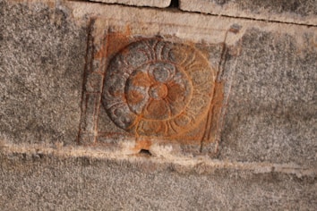 A stone wall features an intricately carved circular pattern with floral and geometric elements, highlighted by a reddish-brown hue. The design is bordered by a square outline, and the stone surface has a rough texture with visible grooves.