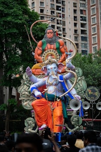 A large, colorful statue of the Hindu deities Hanuman and Ganesha is prominently displayed in an outdoor setting. Ganesha is depicted with a blue complexion and is adorned with vibrant orange and purple garments, decorated with gold jewelry and details. Hanuman is portrayed as muscular and fierce, with an orange hue, sitting behind and slightly above Ganesha. The scene is set amidst a crowded urban environment with residential buildings and lush green trees in the background.