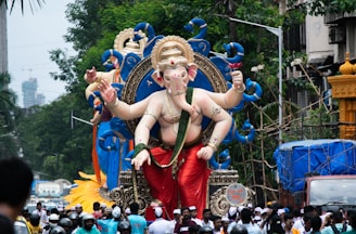 A large, colorful statue of the Hindu deity Ganesha is being paraded through a city street surrounded by a crowd of people. The statue is adorned with ornate decorations, featuring blue peacock motifs and golden details. The street is busy with onlookers and buildings can be seen in the background amidst lush greenery.
