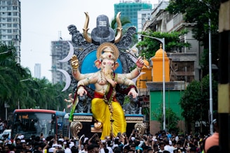A large, ornate statue of the Hindu deity Ganesha is being paraded through a bustling city street. The statue is elaborately decorated with vibrant colors, including yellow, red, and gold, and features the deity's characteristic elephant head and multiple arms. Surrounding the statue is a large crowd of people, some of whom are taking photos and participating in the festival-like atmosphere. The setting includes tall buildings, trees, and a distinct buzz of activity indicative of a celebration.