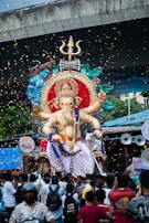 A large statue of the Hindu deity Ganesha is being paraded through a crowd of people. The statue is adorned with vibrant colors, intricate details, and surrounded by floating confetti. The scene is festive, with a blue canopy in the background and a bridge above. Many people are gathered around, taking pictures and participating in the celebration.
