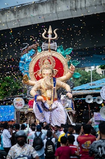 A large statue of the Hindu deity Ganesha is being paraded through a crowd of people. The statue is adorned with vibrant colors, intricate details, and surrounded by floating confetti. The scene is festive, with a blue canopy in the background and a bridge above. Many people are gathered around, taking pictures and participating in the celebration.