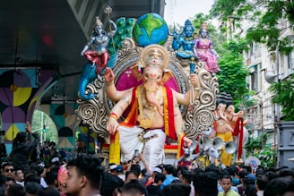 A large, colorful statue of the Hindu deity Ganesha is being paraded through a crowded street. The statue is intricately designed with vibrant colors and detailed ornaments. It is surrounded by numerous people, some of whom are taking photos and others participating in the procession. There are also smaller figures and decorative elements around the main statue, set against a backdrop of urban greenery and buildings.