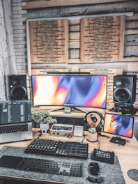 A modern, creative workspace featuring multiple electronic devices. There is an ultrawide monitor with an abstract colorful display, a laptop displaying code or software data, and an additional smaller screen on the right. Below the screens, various audio equipment and a keyboard are organized on a desk, which also holds a few small potted plants. Soundproofing panels are mounted on the wall behind the screens, and large speakers are positioned on each side of the monitor.