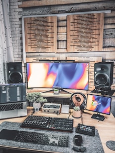 A modern, creative workspace featuring multiple electronic devices. There is an ultrawide monitor with an abstract colorful display, a laptop displaying code or software data, and an additional smaller screen on the right. Below the screens, various audio equipment and a keyboard are organized on a desk, which also holds a few small potted plants. Soundproofing panels are mounted on the wall behind the screens, and large speakers are positioned on each side of the monitor.