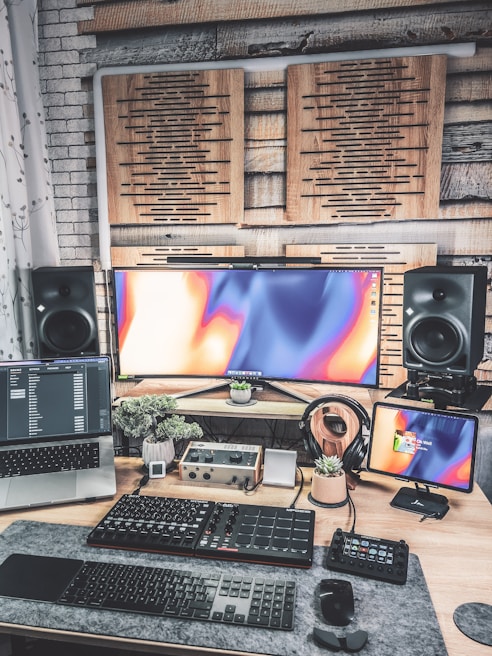A modern, creative workspace featuring multiple electronic devices. There is an ultrawide monitor with an abstract colorful display, a laptop displaying code or software data, and an additional smaller screen on the right. Below the screens, various audio equipment and a keyboard are organized on a desk, which also holds a few small potted plants. Soundproofing panels are mounted on the wall behind the screens, and large speakers are positioned on each side of the monitor.