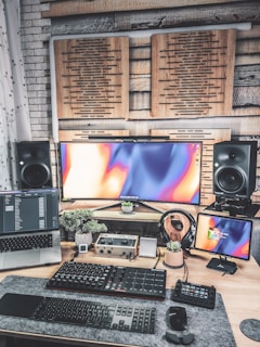 A modern, creative workspace featuring multiple electronic devices. There is an ultrawide monitor with an abstract colorful display, a laptop displaying code or software data, and an additional smaller screen on the right. Below the screens, various audio equipment and a keyboard are organized on a desk, which also holds a few small potted plants. Soundproofing panels are mounted on the wall behind the screens, and large speakers are positioned on each side of the monitor.