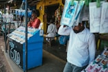 A street vendor stall displaying an array of lottery tickets clipped to a bright blue kiosk. A man in a red shirt leans on the counter, appearing thoughtful, while another man in a white shirt adjusts his hat and walks by. A variety of products hangs from strings above, and a colorful display fills the background with vibrant signs and packets. The scene suggests a busy, lively marketplace.
