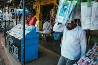 A street vendor stall displaying an array of lottery tickets clipped to a bright blue kiosk. A man in a red shirt leans on the counter, appearing thoughtful, while another man in a white shirt adjusts his hat and walks by. A variety of products hangs from strings above, and a colorful display fills the background with vibrant signs and packets. The scene suggests a busy, lively marketplace.