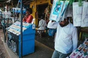 A street vendor stall displaying an array of lottery tickets clipped to a bright blue kiosk. A man in a red shirt leans on the counter, appearing thoughtful, while another man in a white shirt adjusts his hat and walks by. A variety of products hangs from strings above, and a colorful display fills the background with vibrant signs and packets. The scene suggests a busy, lively marketplace.