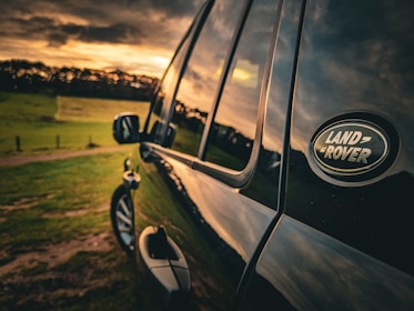 A close-up view of the side of a Land Rover vehicle, with a clear focus on the emblem. The vehicle is parked in a grassy field during sunset, casting warm golden hues onto the car's surface. The background features an open landscape with a few trees silhouetted against the sky, creating a serene and picturesque setting.