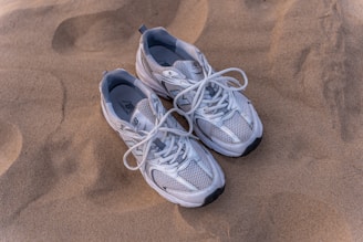 A pair of white and gray sneakers with white laces are lying on a sandy surface. The sneakers have a mesh design which suggests they are breathable and lightweight.