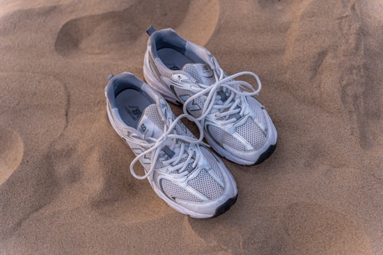 A pair of white and gray sneakers with white laces are lying on a sandy surface. The sneakers have a mesh design which suggests they are breathable and lightweight.