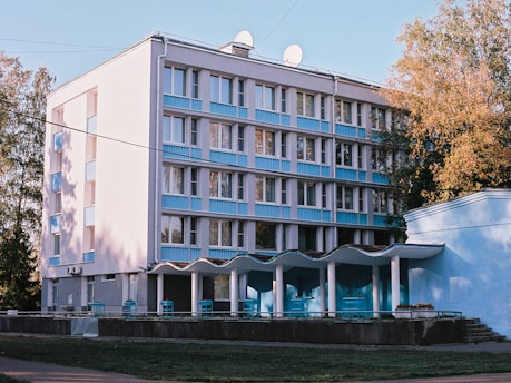 A modern, rectangular building with several floors and a facade featuring blue and white colors. There are multiple windows and balconies, and two satellite dishes are visible on the roof. The building is adjacent to lush trees, indicating an urban environment mixed with greenery. In front of the building, there is a covered entrance with a wavy roof supported by columns.