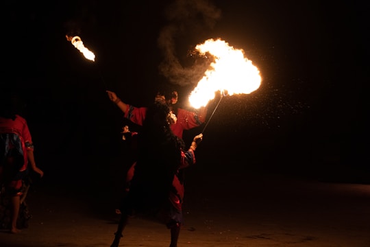 A performer engages in a fire dance at night. The individual, dressed in colorful attire, holds a flaming torch, creating mesmerizing patterns in the dark. The background is completely black, highlighting the brightness and intensity of the flames.