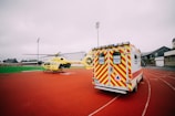 A yellow emergency helicopter and a white ambulance with yellow and red stripes are parked on an outdoor track. The background features overcast skies and buildings.