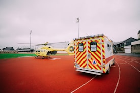A yellow emergency helicopter and a white ambulance with yellow and red stripes are parked on an outdoor track. The background features overcast skies and buildings.
